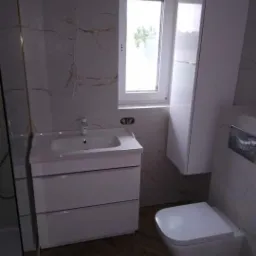 Nowoczesna łazienka z białymi meblami, marmurowymi płytkami i drewnianą podłogą w jodełkę, widok na wiszącą szafkę i toaletę.