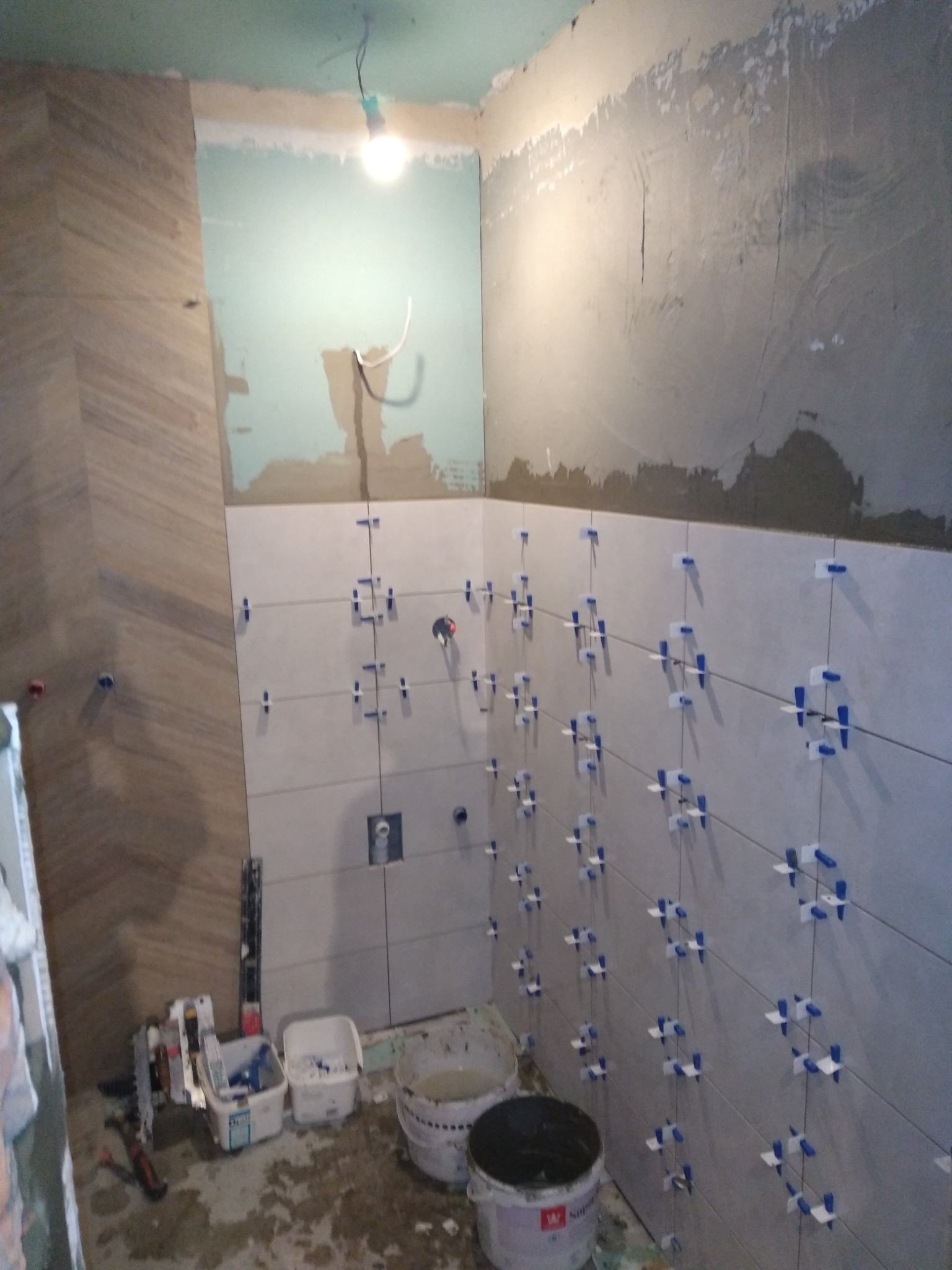 Remont łazienki w trakcie: białe płytki z niebieskimi krzyżykami dystansowymi na ścianie, częściowo wykończona ściana z płytkami imitującymi drewno, widoczne narzędzia i wiadra z zaprawą.