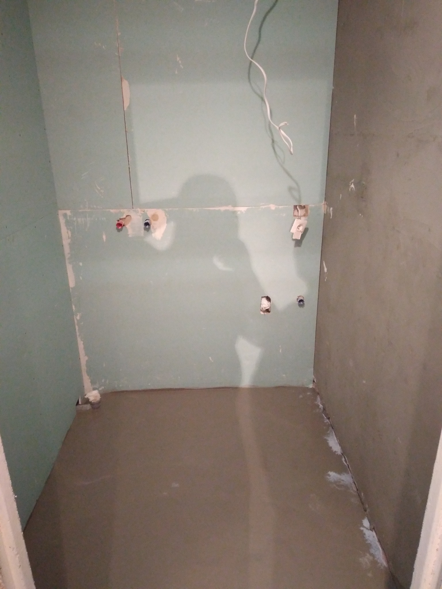 Surowy stan łazienki w trakcie prac: ściany pokryte płytami kartonowo-gipsowymi w odcieniach zieleni i szarości, widoczne instalacje wodne i elektryczne, wylewka podłogowa, cień osoby...