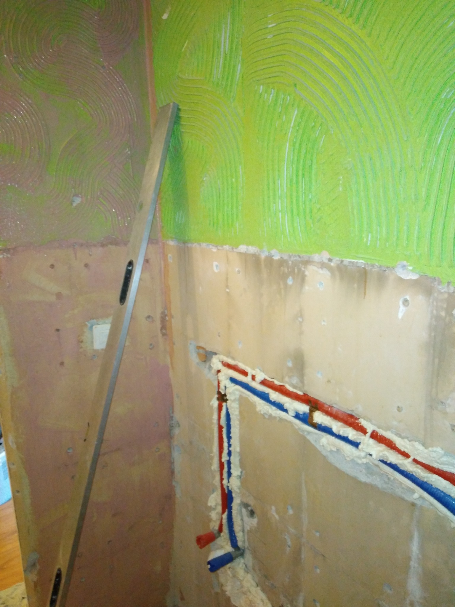 Ściana w trakcie przygotowywania do położenia płytek, widoczne ślady po kleju, poziomica oparta o ścianę, wystające rury instalacji wodnej zabezpieczone pianką.