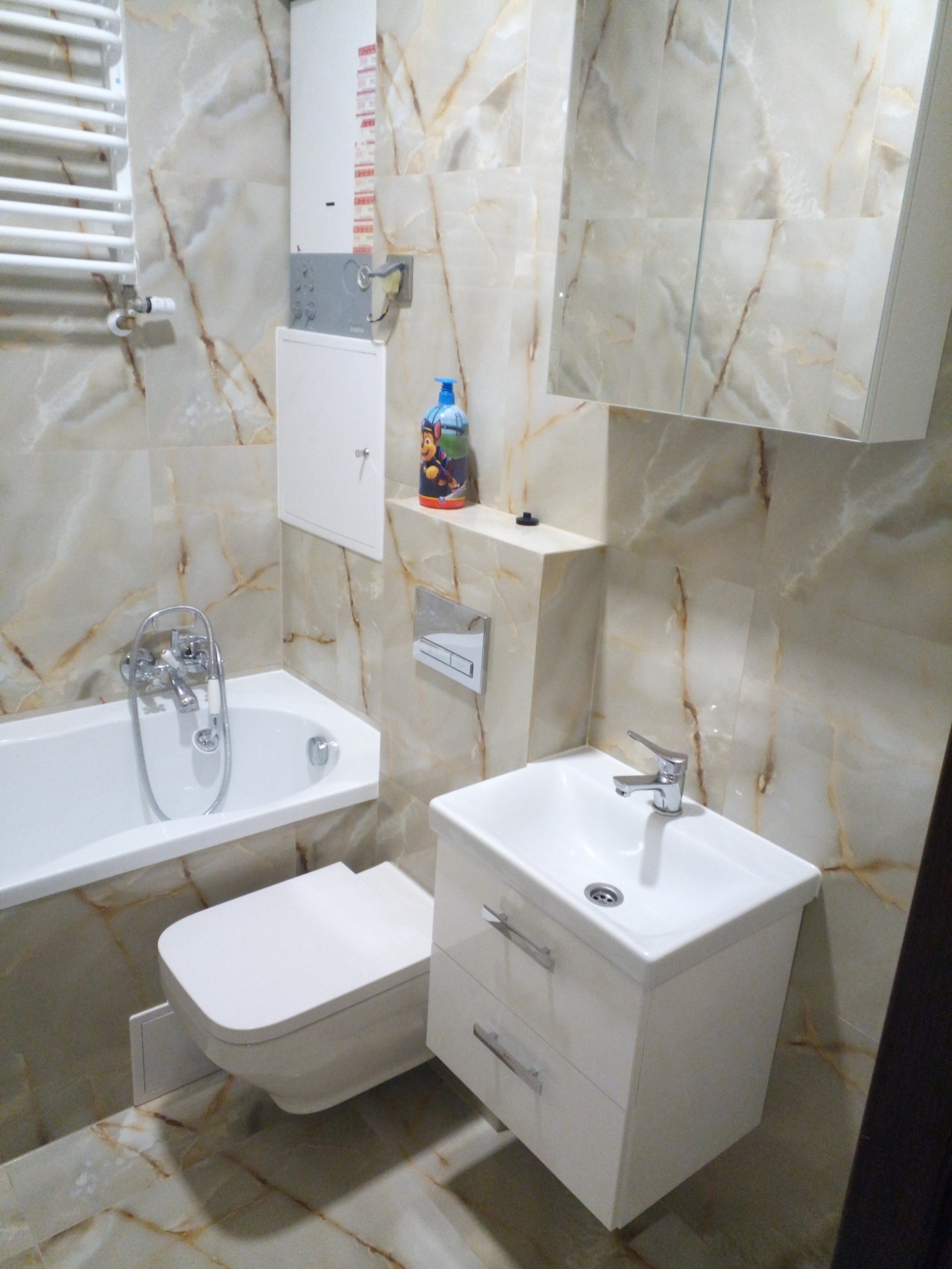 Wykończona łazienka z beżowymi płytkami imitującymi marmur, biała wanna z chromowaną baterią, podwieszana toaleta, umywalka z szafką i lustrzana szafka wisząca, na półce kolorowa butelka z płynem...