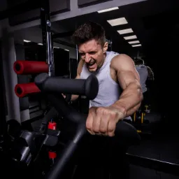 Mężczyzna z zaciśniętymi pięściami, z intensywnym wyrazem twarzy, ćwiczy na maszynie w siłowni, widoczny w lustrze drugi mężczyzna w tle.