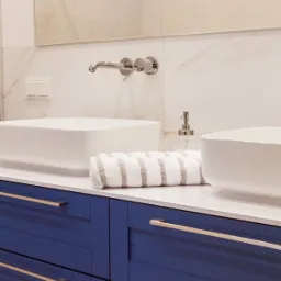 Nowoczesna łazienka z dwoma białymi umywalkami nablatowymi, bateria ścienna, niebieska szafka z poziomymi uchwytami i zwinięty ręcznik w paski.