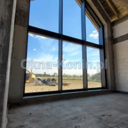 Wnętrze w stanie surowym z zamontowanym dużym, aluminiowym oknem trapezowym w kolorze antracytowym, przez które widać błękitne niebo i sprzęt budowlany na zewnątrz.