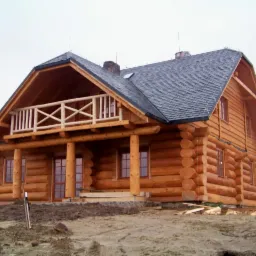 Gotowy dom z okrąglaków w stylu kanadyjskim z balkonem, pokryty szarą dachówką, na tle pochmurnego nieba i fragmentu lasu.