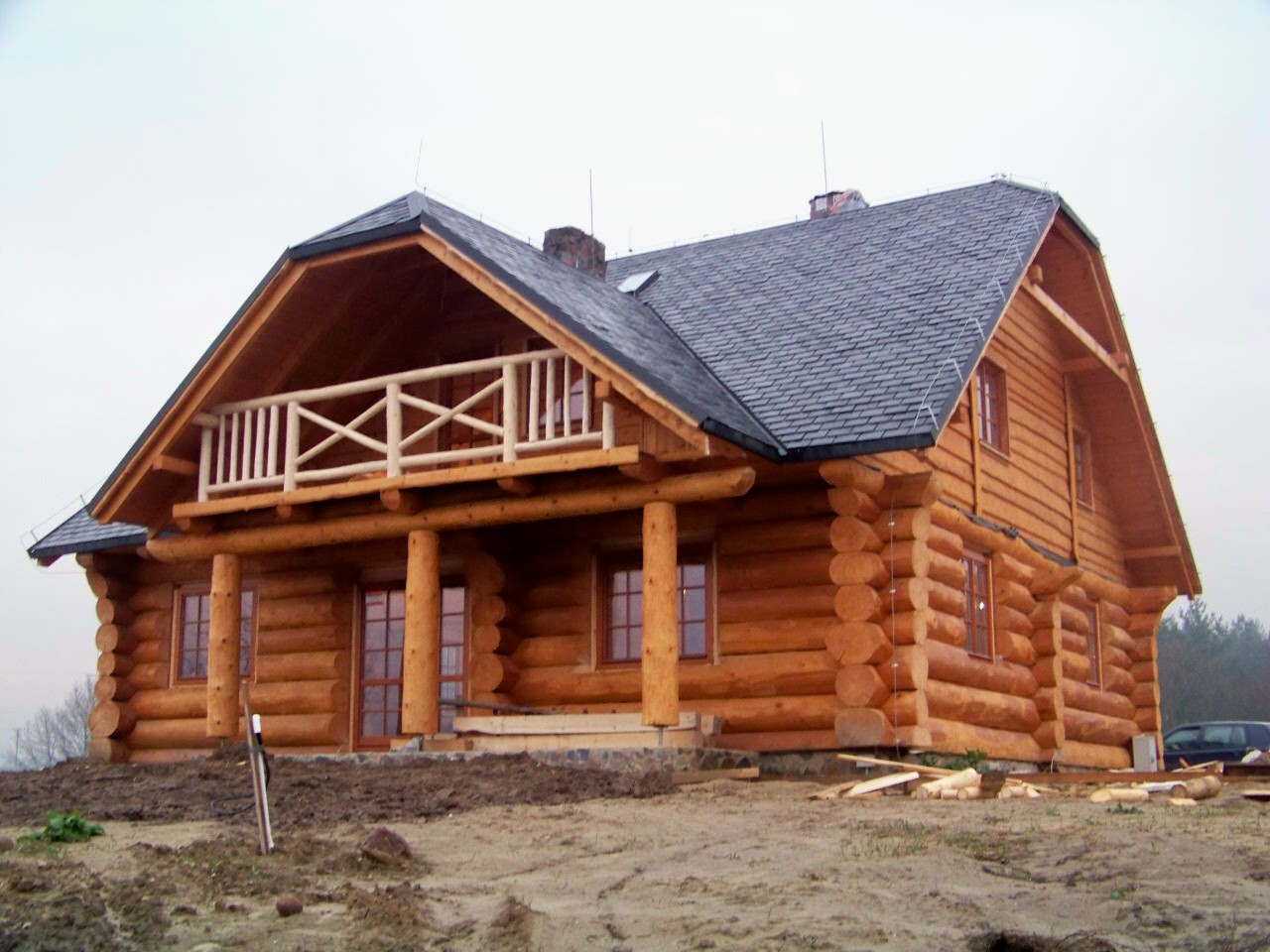 Gotowy dom z okrąglaków w stylu kanadyjskim z balkonem, pokryty szarą dachówką, na tle pochmurnego nieba i fragmentu lasu.