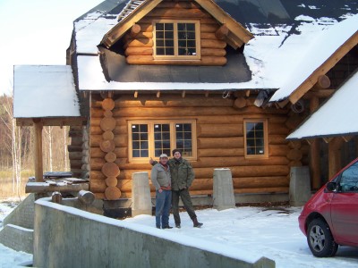 Dwaj mężczyźni stoją przed częściowo ośnieżonym domem z okrąglaków, z widocznym fragmentem czerwonego samochodu po prawej stronie kadru, na tle zimowego krajobrazu.