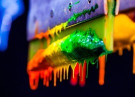 Rakla z resztkami farb w kolorach tęczy, ściekających kroplami, używana w procesie sitodruku, widoczna z bliska na ciemnym tle.