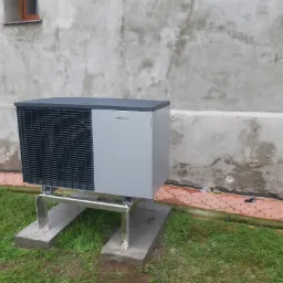 Szara pompa ciepła Viessmann Vitocal 200, zamontowana na metalowej konstrukcji i betonowych podstawach, ustawiona na trawie przy szarej ścianie budynku z małym oknem