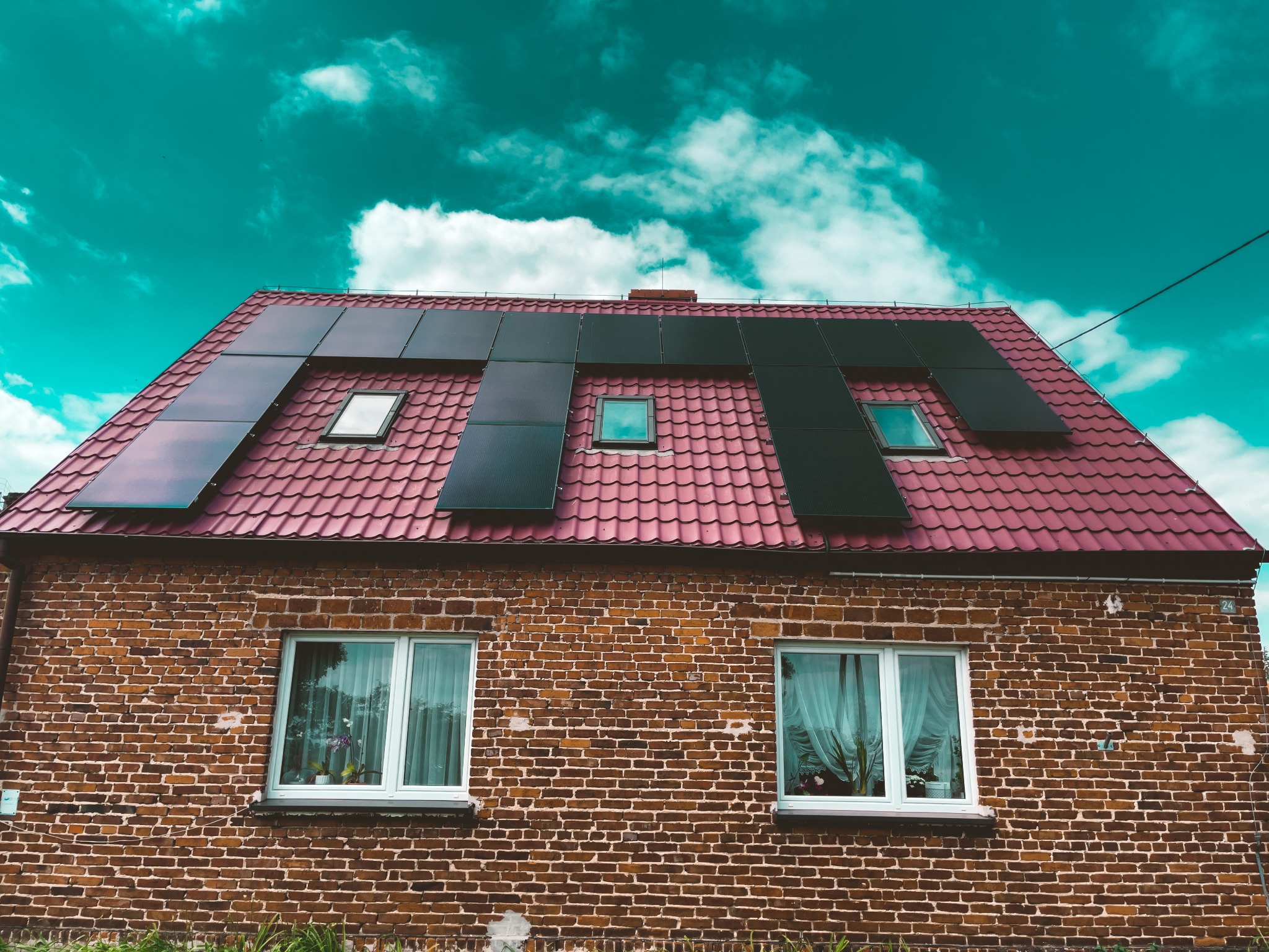 Czerwony dach z zamontowanymi panelami słonecznymi na ceglanym domu jednorodzinnym z dwoma oknami, na tle błękitnego nieba z chmurami.