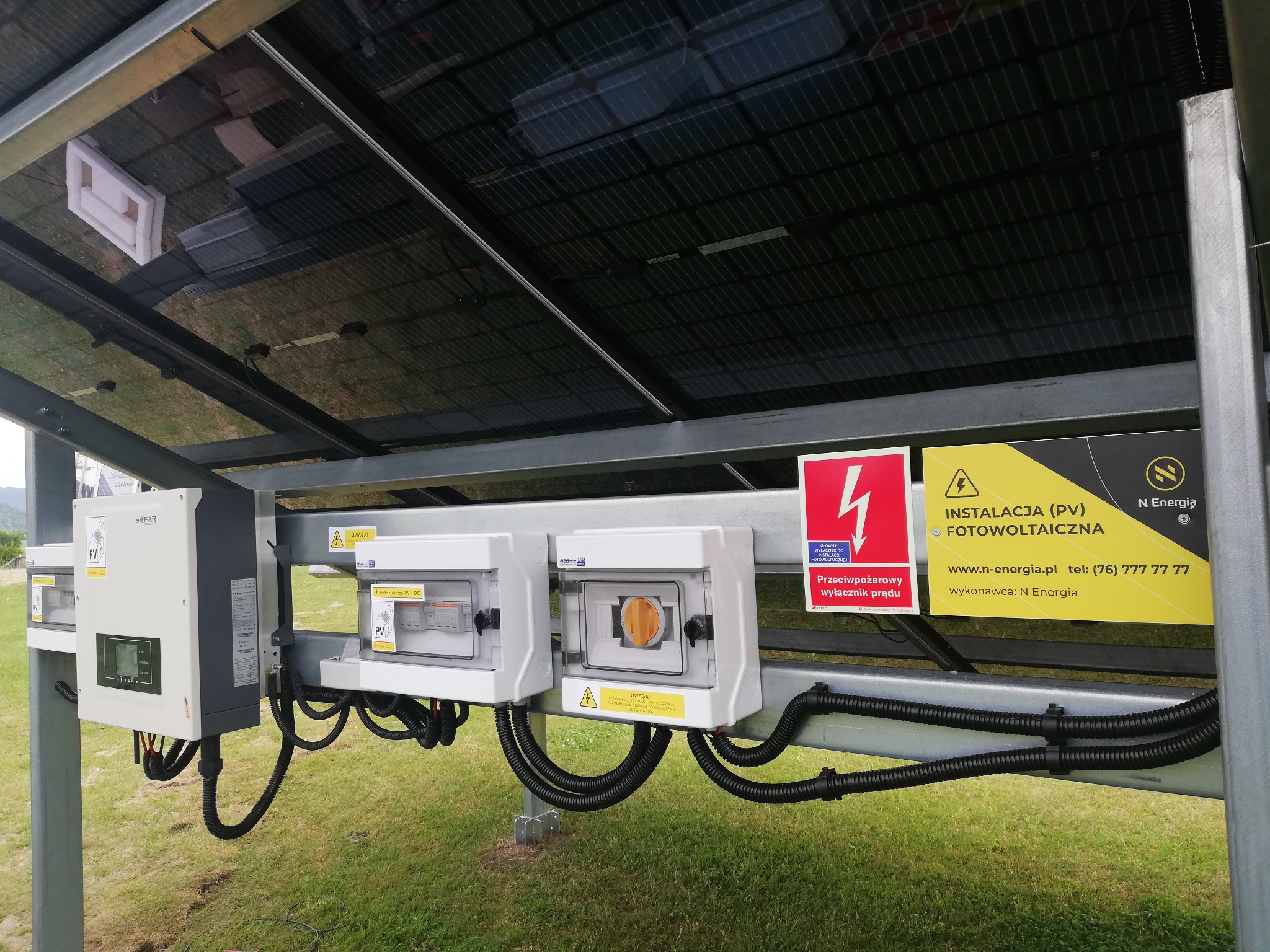 Instalacja paneli słonecznych z widocznymi elementami elektrycznymi, rozdzielnicami i okablowaniem; tabliczka informacyjna z logo firmy.