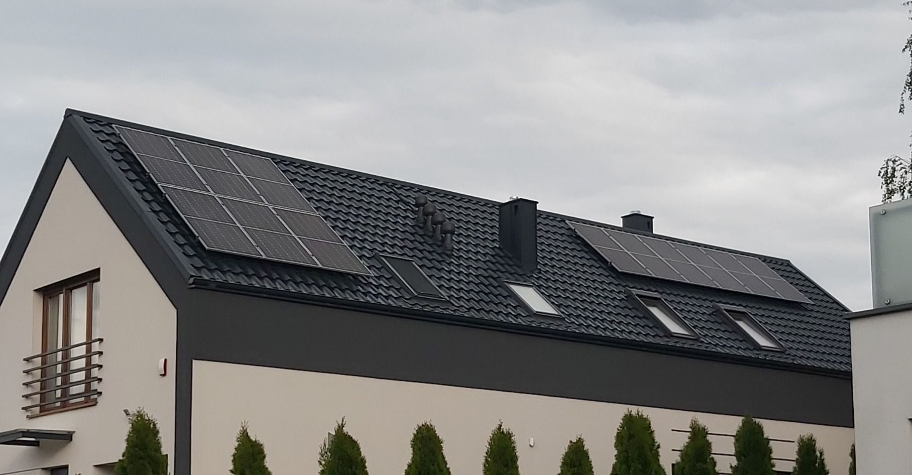 Nowoczesny dom z ciemną dachówką, oknami dachowymi i panelami słonecznymi na dachu, widok z poziomu gruntu w pochmurny dzień.