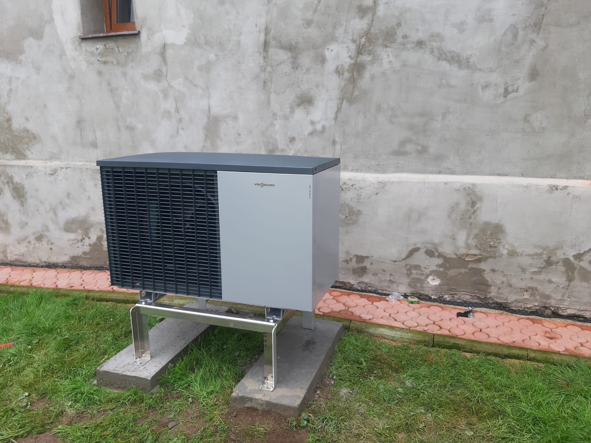 Szara pompa ciepła Viessmann Vitocal 200, zamontowana na metalowej konstrukcji i betonowych podstawach, ustawiona na trawie przy szarej ścianie budynku z małym oknem