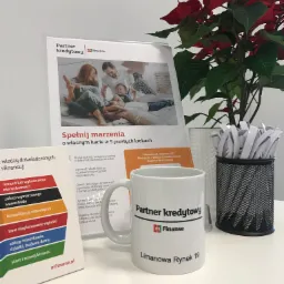 Biurko z kubkiem i materiałami informacyjnymi Partner Kredytowy mFinanse, w tle poinsecja i pojemnik z długopisami.