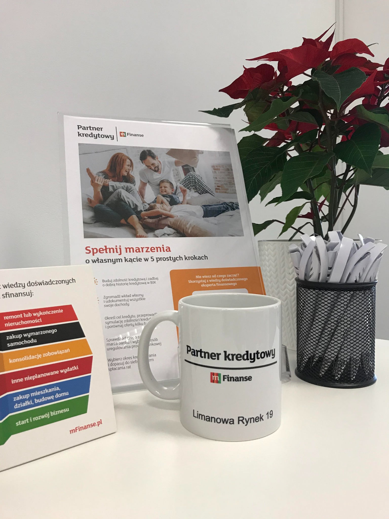 Biurko z kubkiem i materiałami informacyjnymi Partner Kredytowy mFinanse, w tle poinsecja i pojemnik z długopisami.