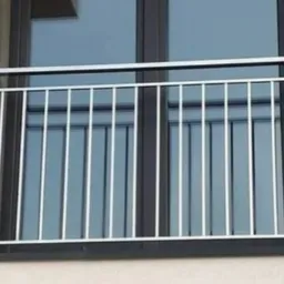 Nierdzewna balustrada balkonowa z pionowymi prętami, zamontowana na tle okna z ciemną ramą i szarą elewacją.