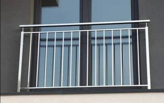 Nierdzewna balustrada balkonowa z pionowymi prętami, zamontowana na tle okna z ciemną ramą i szarą elewacją.