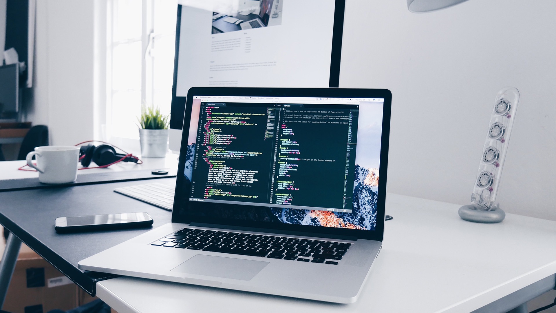 Laptop z otwartym edytorem kodu na biurku, obok drugi monitor, telefon, słuchawki i kubek, roślina w doniczce w tle, minimalistyczne biuro.