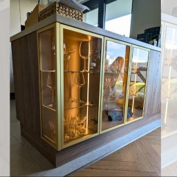 EUDI DESIGN VOLHA SHURPITSKAYA - Drewniana witryna z przeszklonymi drzwiami i złotymi ramkami, podświetlona od wewnątrz, eksponująca kolekcję szkła. Widoczny parkiet na podłodze i jasne tło.