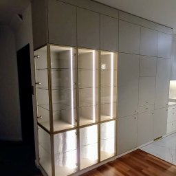 EUDI DESIGN VOLHA SHURPITSKAYA - Elegancka zabudowa meblowa w salonie z podświetlanymi witrynami i frontami z ryflowanego szkła, uzupełniona o szafki i szuflady w odcieniach beżu. Parkiet na podłodze.