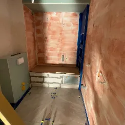 Łazienka w trakcie remontu: Ściany przygotowane pod płytki, podłoga wyłożona płytkami, widoczne krzyżyki dystansowe, wnęka z półką w trakcie wykańczania, fragment poziomicy w rogu.