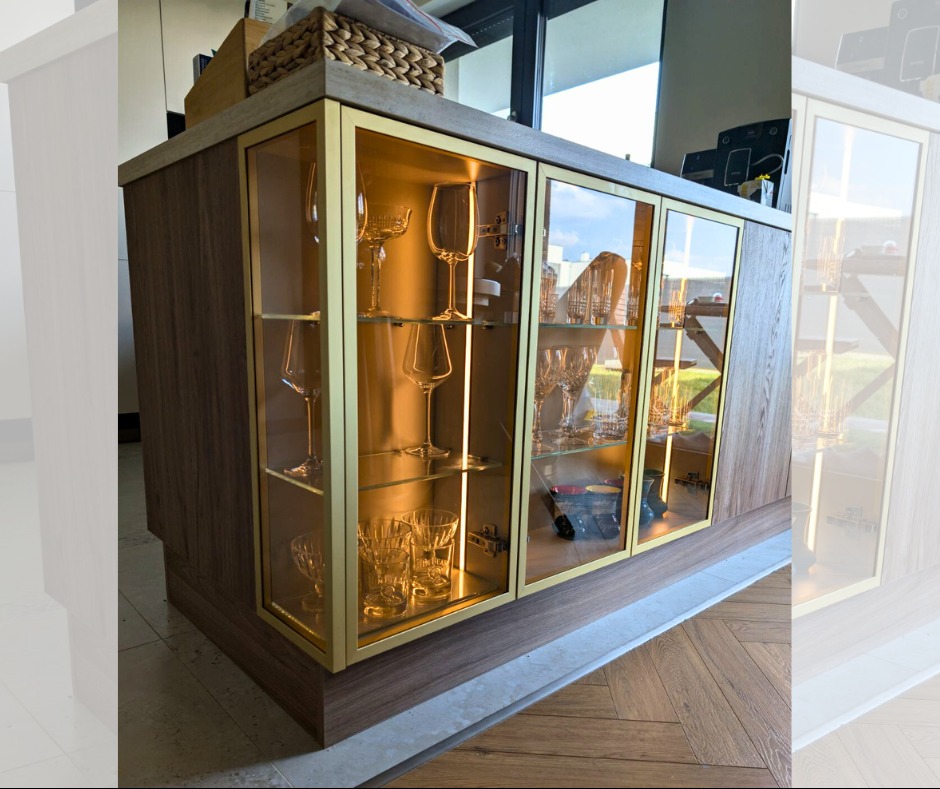 Drewniana witryna z przeszklonymi drzwiami i złotymi ramkami, podświetlona od wewnątrz, eksponująca kolekcję szkła. Widoczny parkiet na podłodze i jasne tło.