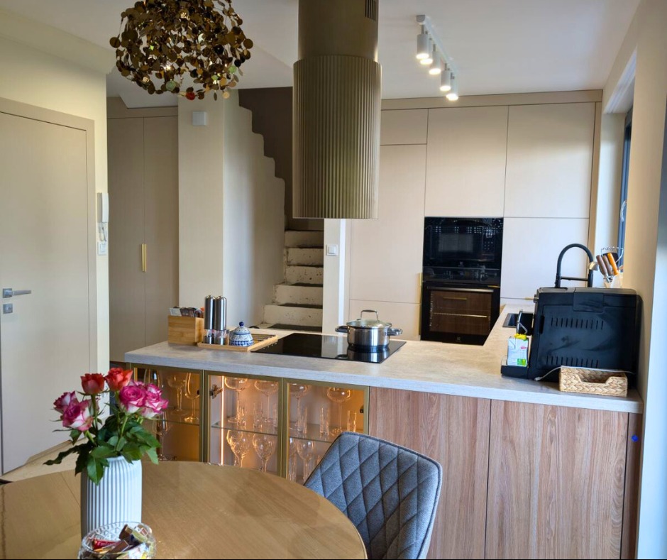 Beżowa kuchnia z drewnianymi akcentami: wyspa z podświetlanymi szafkami na kieliszki, okrągły stół z różami, schody w tle, nowoczesny design.