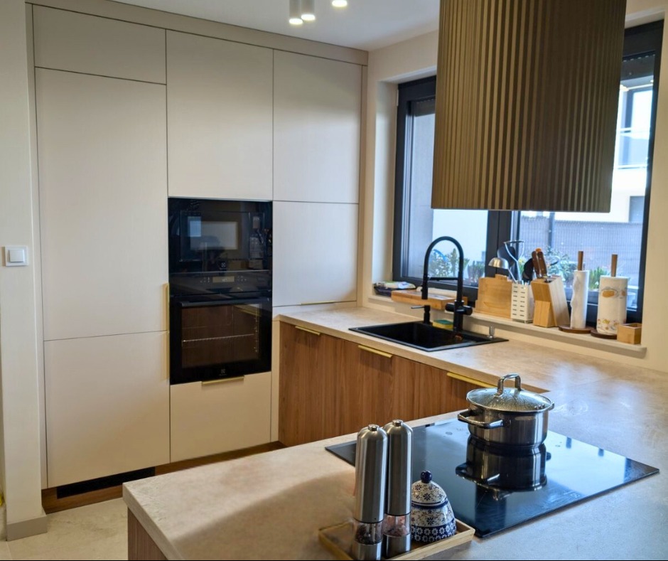 Nowoczesna kuchnia z zabudową w odcieniach bieli i drewna. Widoczny piekarnik, płyta indukcyjna z garnkiem, czarny zlew i okno z widokiem na zewnątrz.