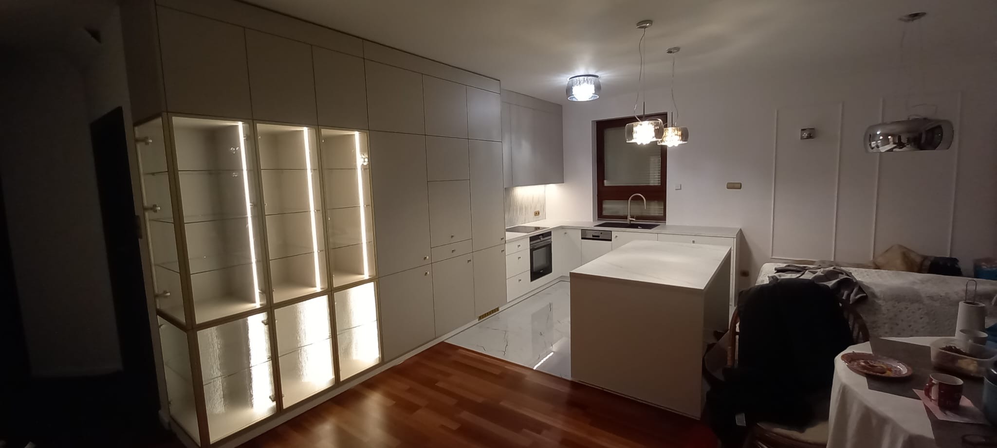 Nowoczesna kuchnia z zabudową meblową w odcieniach bieli i szarości, z wyspą i oświetleniem LED w witrynach. Parkiet w ciepłym odcieniu brązu.