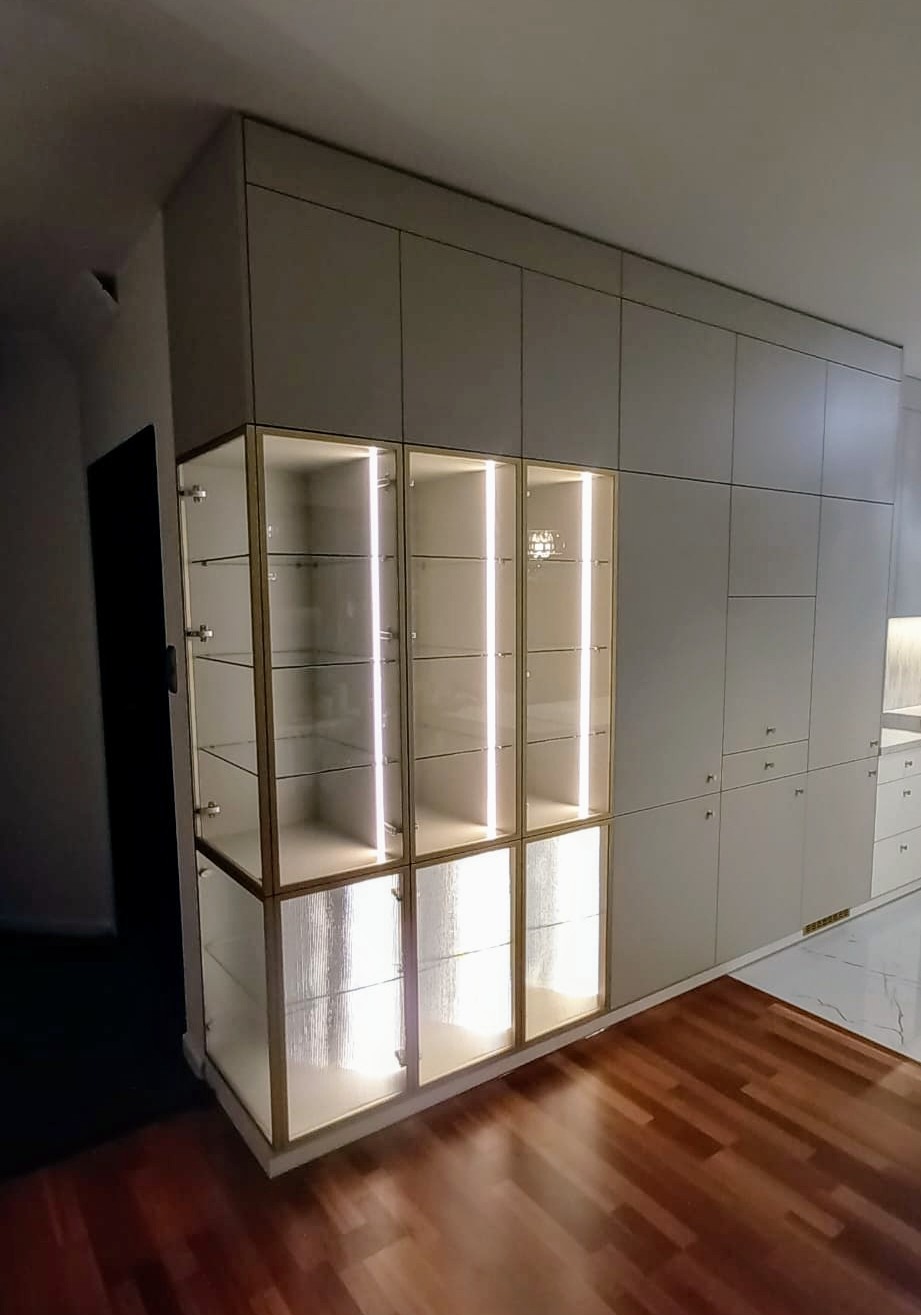Elegancka zabudowa meblowa w salonie z podświetlanymi witrynami i frontami z ryflowanego szkła, uzupełniona o szafki i szuflady w odcieniach beżu. Parkiet na podłodze.