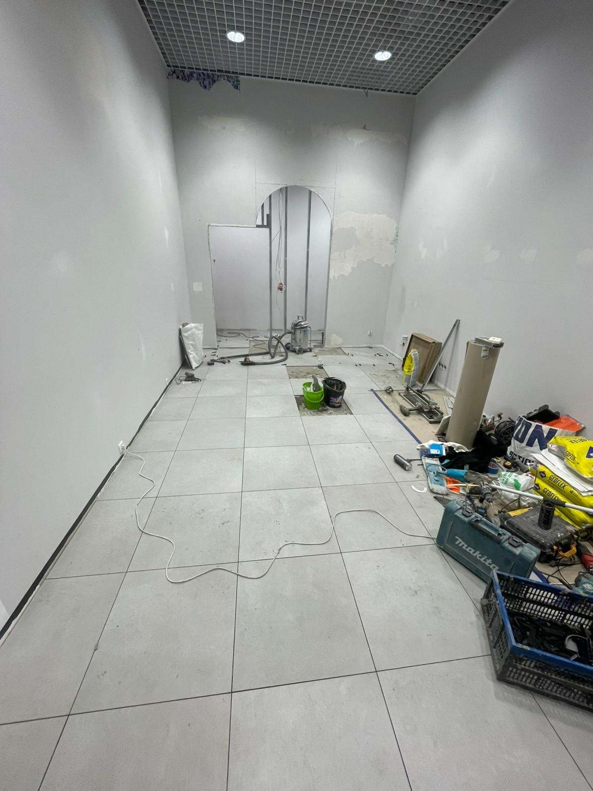 Wnętrze w trakcie remontu: ułożone szare płytki podłogowe, ściany przygotowane do malowania, widoczna konstrukcja łukowego otworu drzwiowego i narzędzia glazurnicze.