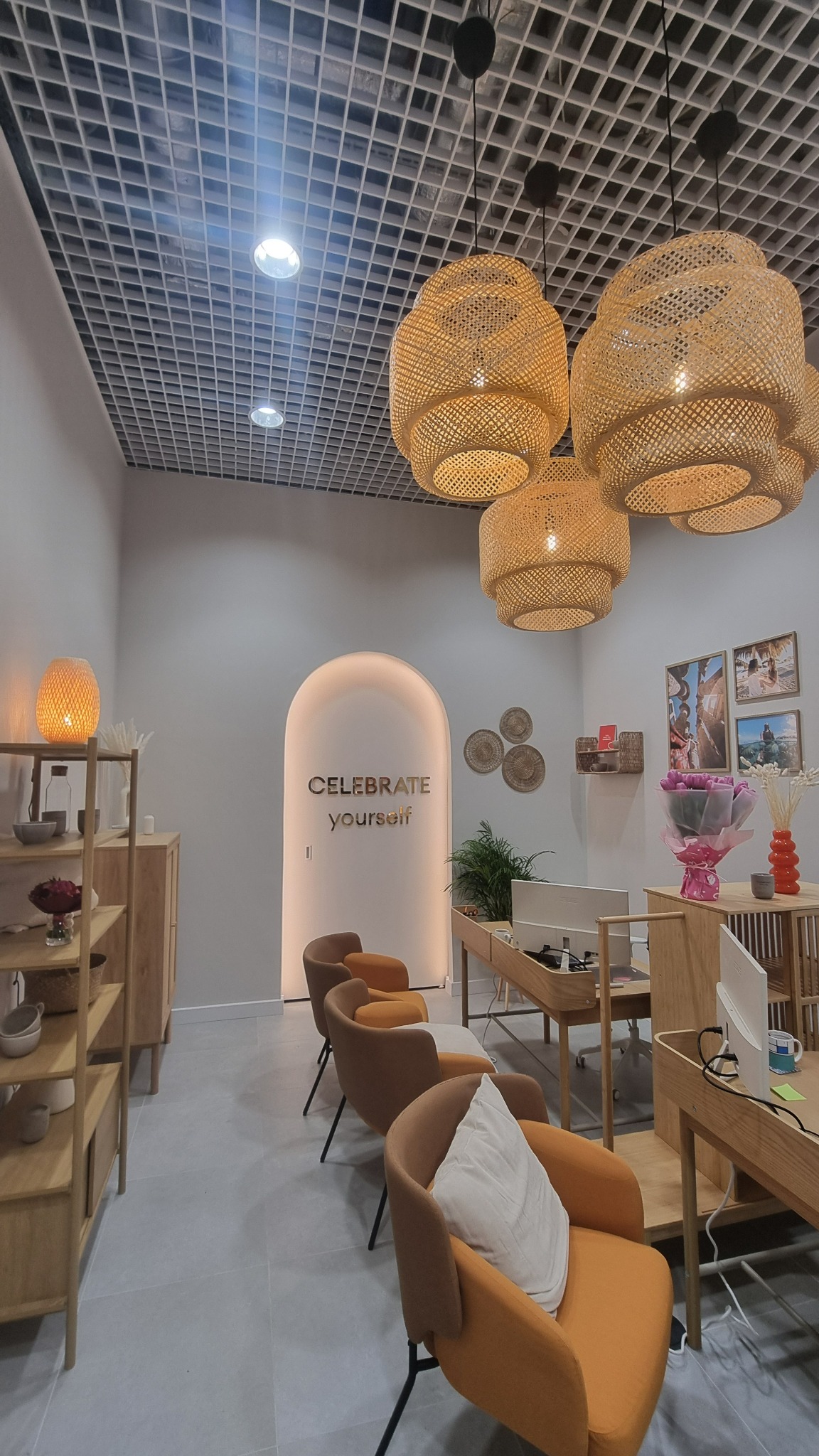Jasne wnętrze biura z rattanowymi lampami, drewnianymi meblami i szarą podłogą. W tle łuk z napisem 'Celebrate Yourself'.