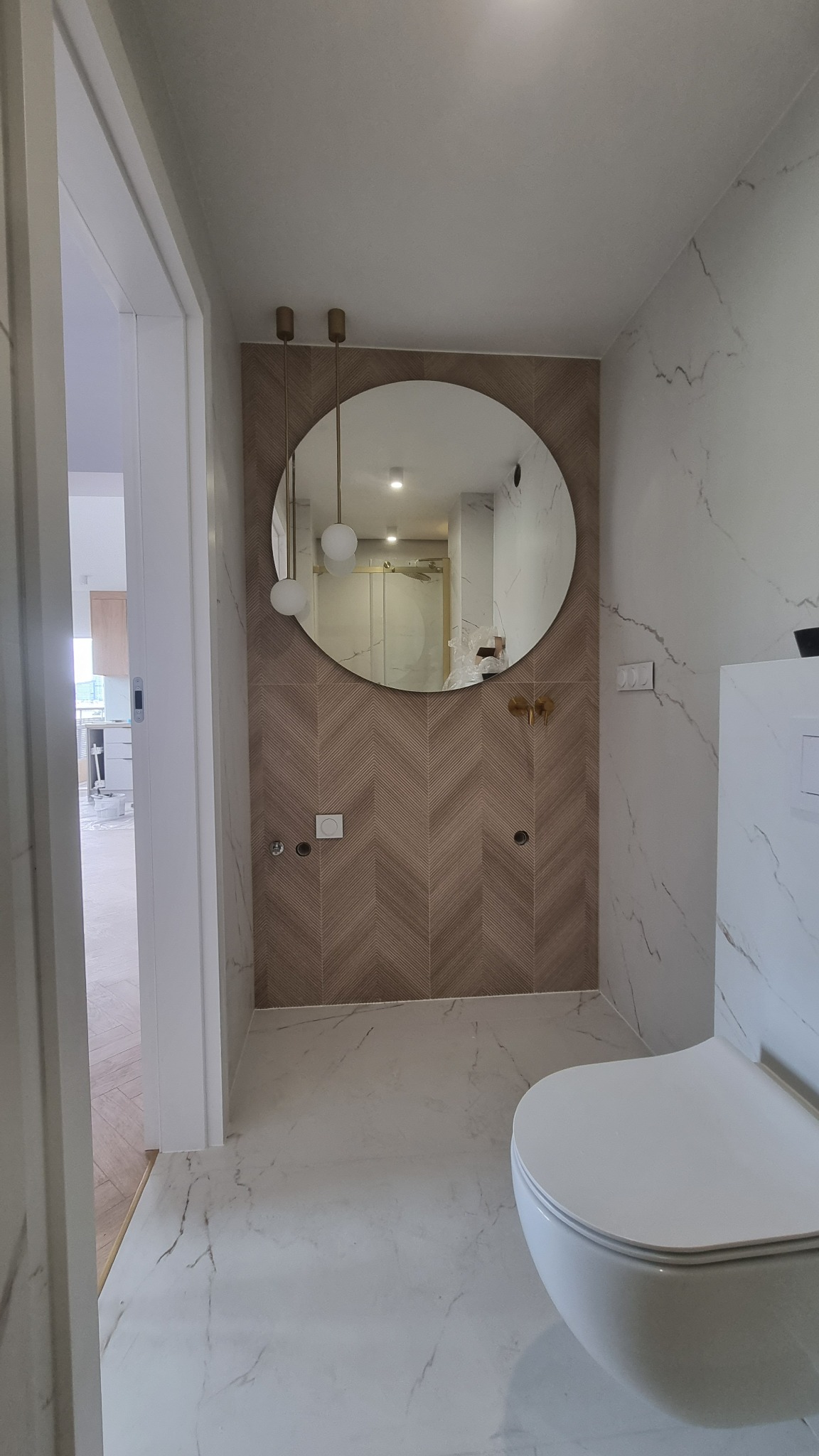 Nowoczesna łazienka z okrągłym lustrem i wzorem jodełki na ścianie. Biała ceramika i marmurowe płytki podłogowe. Widok zza drzwi.