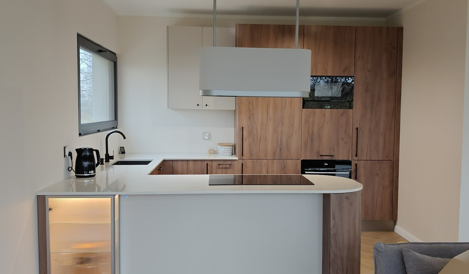 Nowoczesna kuchnia z wyspą: białe blaty, drewniane szafki, czarne akcenty. Minimalistyczny design, oświetlenie LED, okno z czarną ramą. Widok na wnętrze.