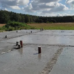 Wylany fundament z wystającymi rurami kanalizacyjnymi, widok na łąkę i las w tle, słoneczny dzień.