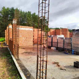 Budowa domu z czerwonej cegły: widoczne zbrojenia pionowe, częściowo wzniesione mury, palety z cegłami w folii w tle, teren budowy z zielenią i lasem w tle.