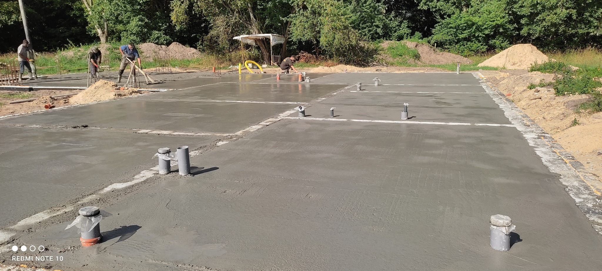 Wylany fundament z wystającymi rurami kanalizacyjnymi, w tle pracujący robotnicy i pryzmy piasku.