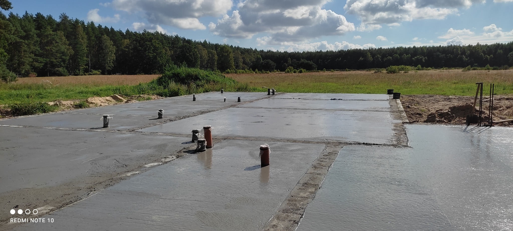 Wylany fundament z wystającymi rurami kanalizacyjnymi, widok na łąkę i las w tle, słoneczny dzień.