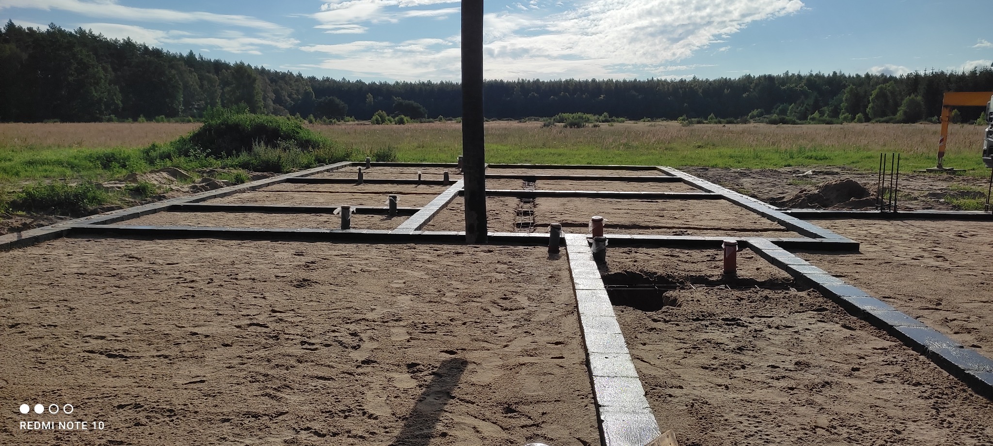 Przygotowanie fundamentów: zarys fundamentu z ułożonych bloczków betonowych, widoczne pionowe rury instalacyjne, w tle łąka i las.