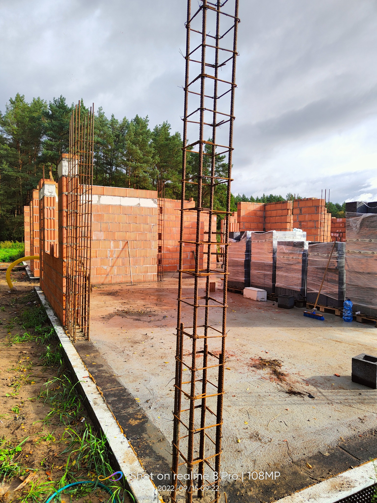 Budowa domu z czerwonej cegły: widoczne zbrojenia pionowe, częściowo wzniesione mury, palety z cegłami w folii w tle, teren budowy z zielenią i lasem w tle.
