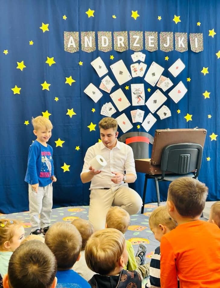 Iluzjonista w białej koszuli prezentuje trik z rolką papieru toaletowego przed grupą dzieci na tle dekoracji andrzejkowej z gwiazdami i kartami.