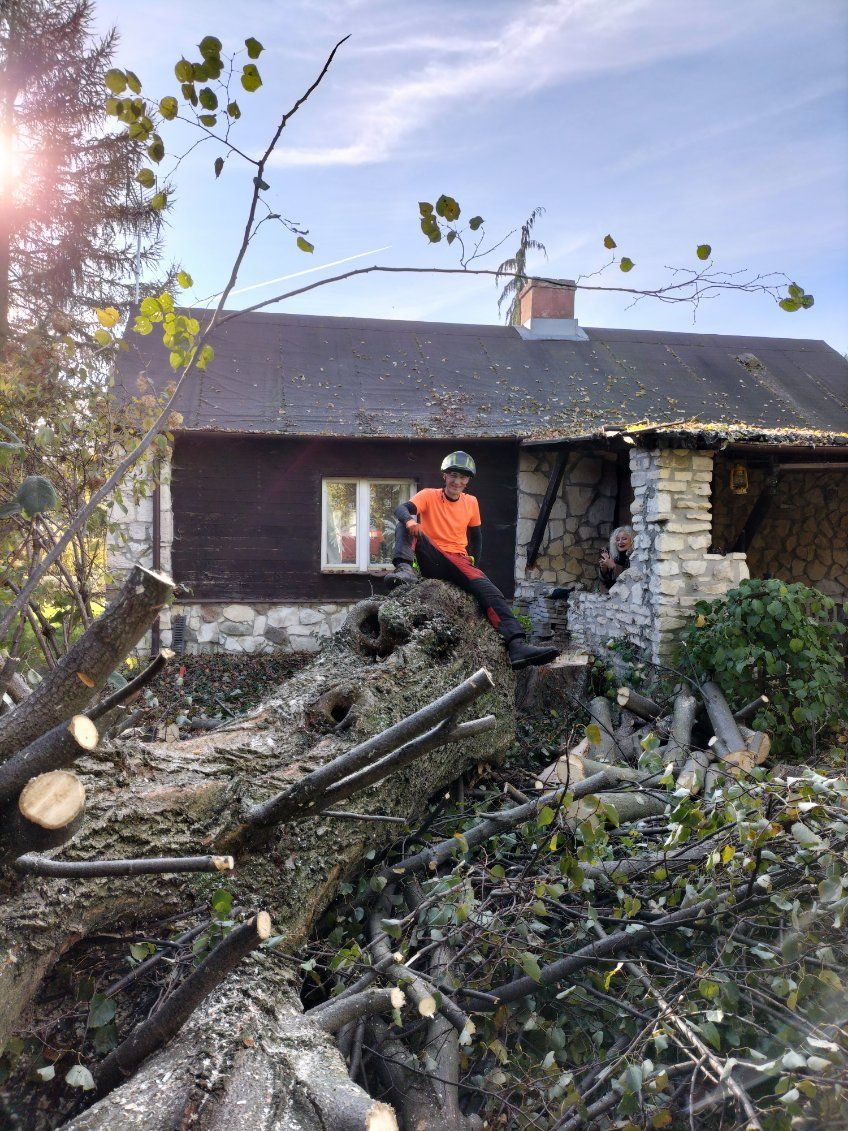 Mężczyzna w kasku i pomarańczowej koszulce siedzi na powalonym pniu drzewa przed domem z kamienną elewacją, w tle widać osobę wyglądającą z okna