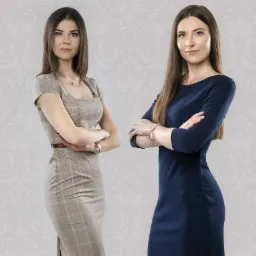 Portret dwóch kobiet w sukienkach biznesowych na tle wzorzystej ściany, z założonymi rękami i poważnym wyrazem twarzy.