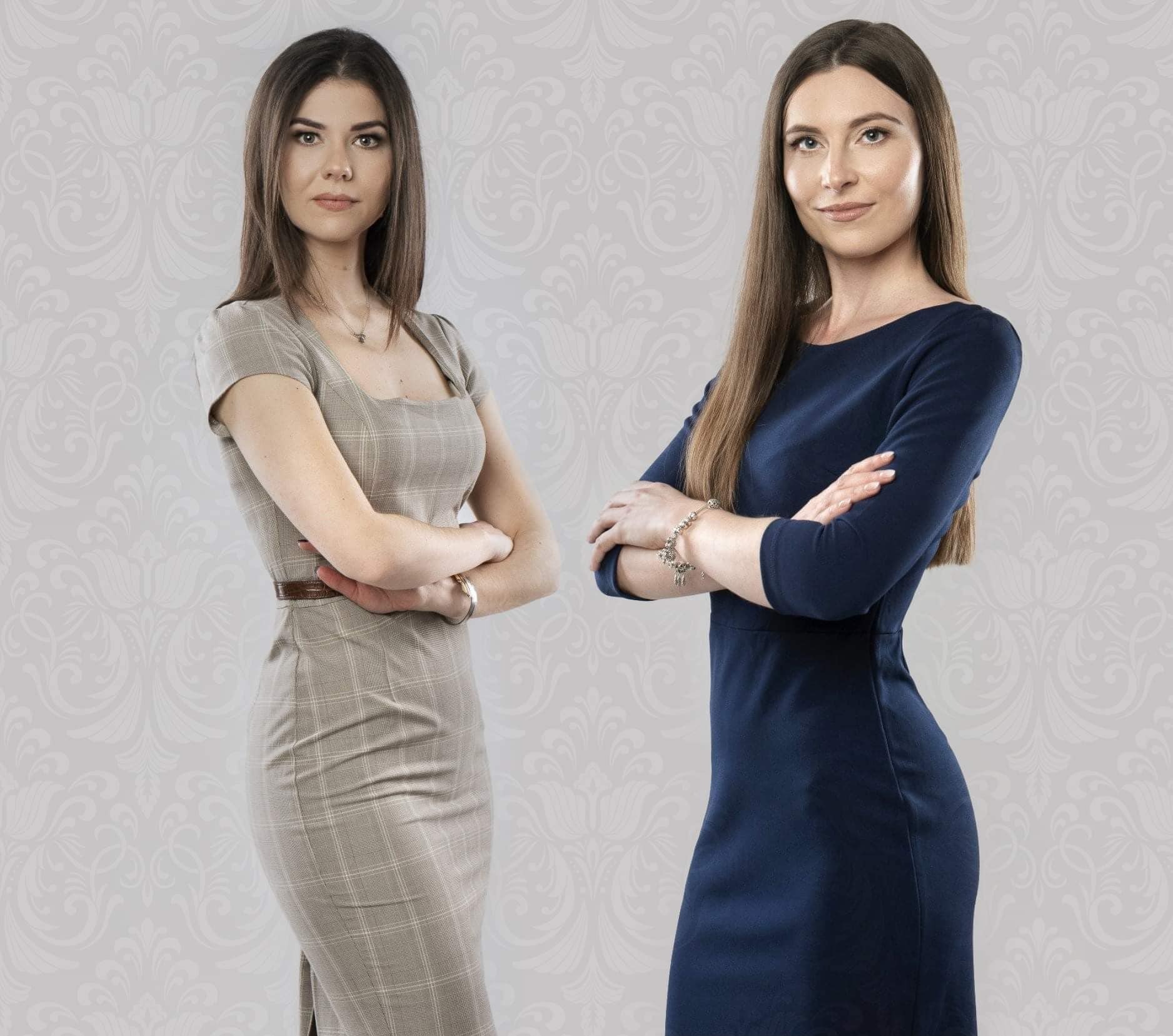 Portret dwóch kobiet w sukienkach biznesowych na tle wzorzystej ściany, z założonymi rękami i poważnym wyrazem twarzy.