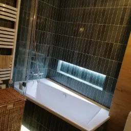 Nowoczesna łazienka z wanną obudowaną ciemnymi, wąskimi płytkami ułożonymi poziomo, podświetlona listwą LED. Nad wanną przeszklony parawan. W ścianie wnęka z oświetleniem.