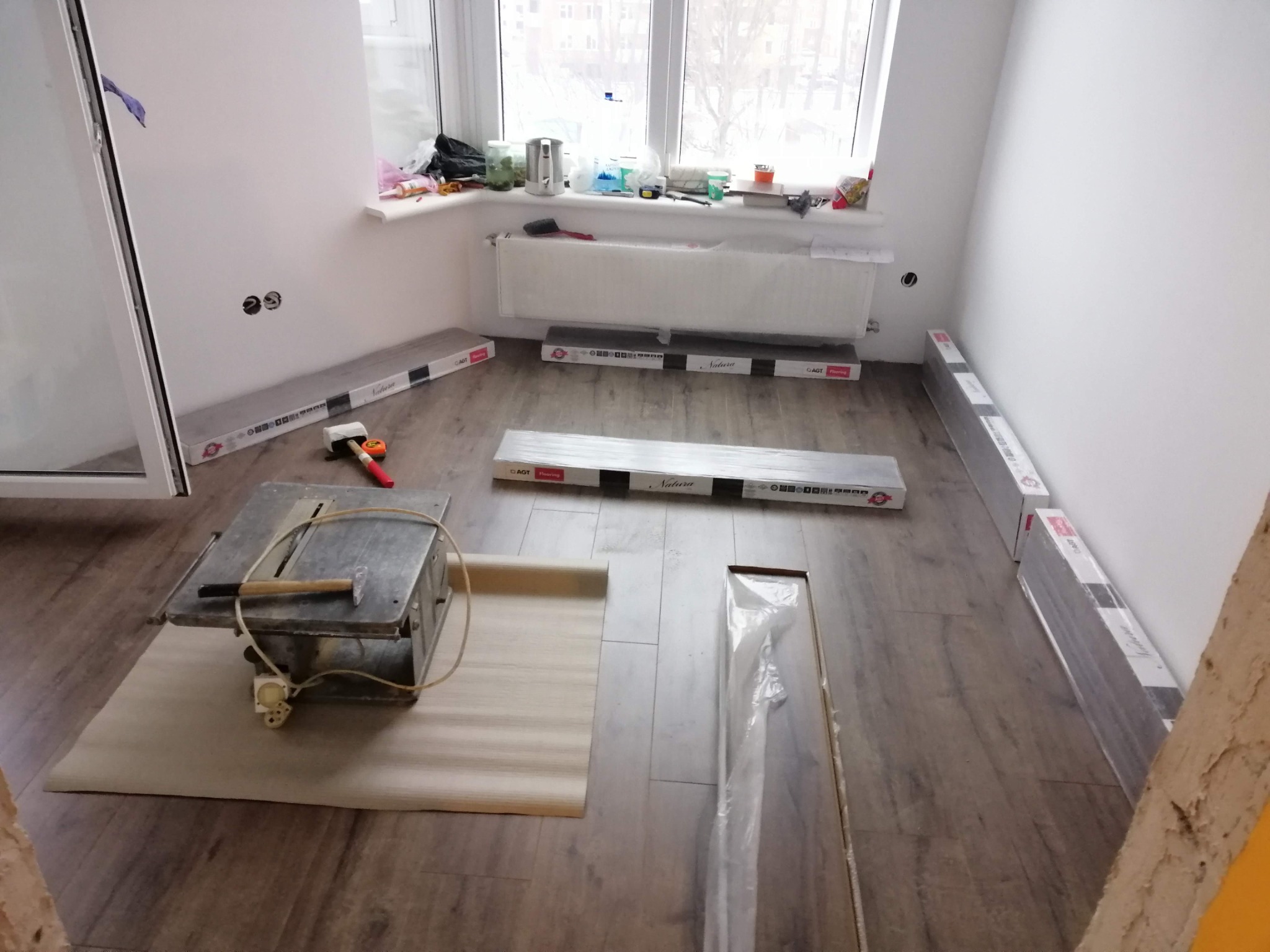 Przygotowania do montażu paneli podłogowych: piła stołowa, paczki paneli AGT Natura, młotek gumowy i podkład w pokoju z oknem.