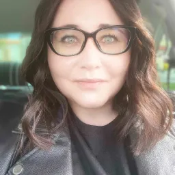 Portret kobiety w czarnej skórzanej kurtce i okularach o kocim kształcie, z delikatnym uśmiechem, zrobiony w samochodzie w słoneczny dzień.