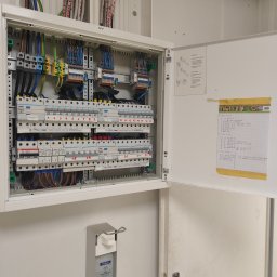 Instalacje Elektryczne - Otwarta skrzynka rozdzielcza z bezpiecznikami i okablowaniem, z naklejoną kartką z opisem obwodów, oraz dozownik do dezynfekcji rąk na ścianie poniżej.
