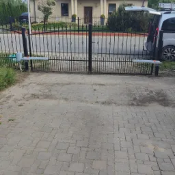 Czarna, metalowa brama wjazdowa z automatyką, widok na dom z beżową elewacją i szarym dachem, podjazd z kostki brukowej.