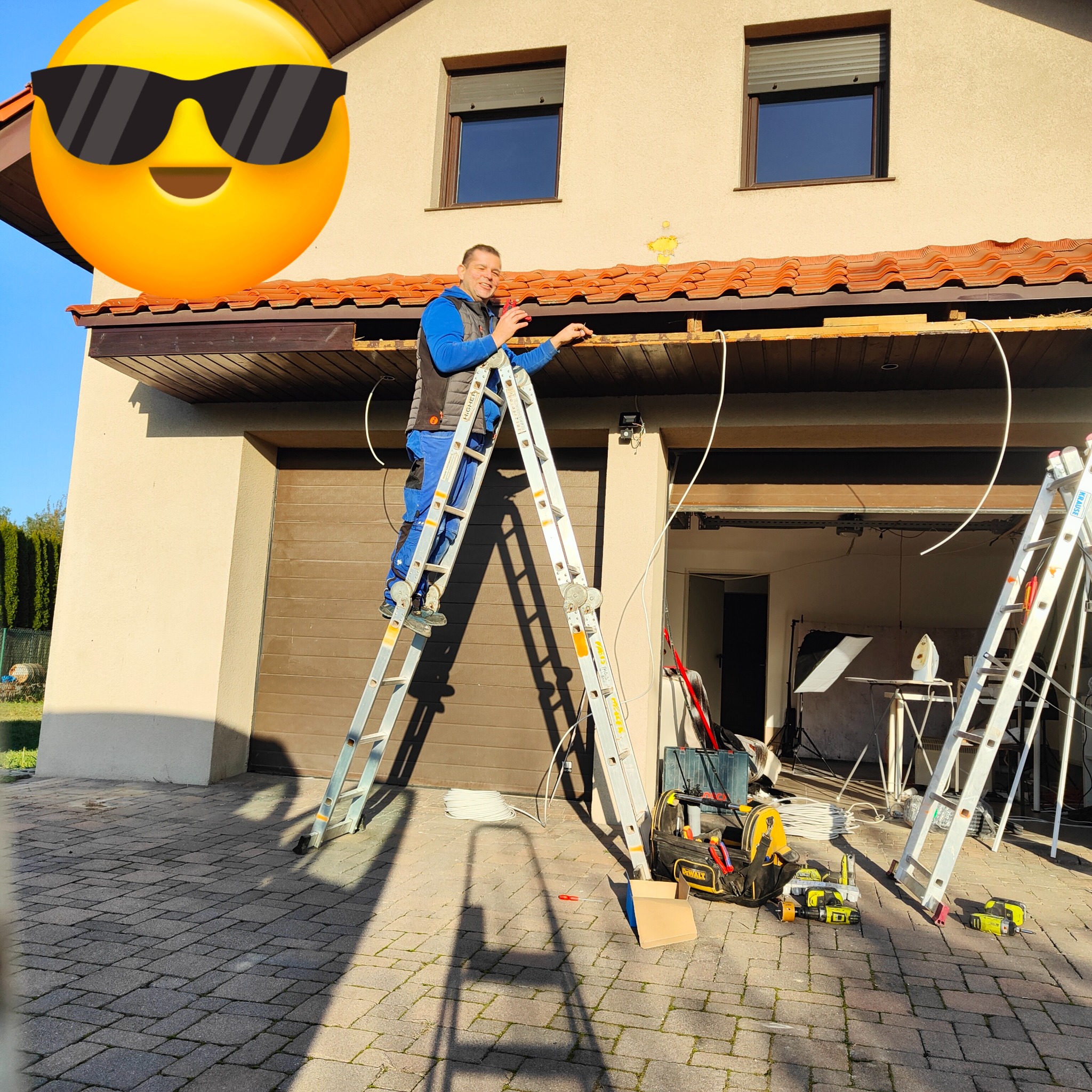 Elektryk na drabinie montuje przewody pod okapem domu jednorodzinnego, narzędzia i skrzynka leżą na brukowanym podjeździe.